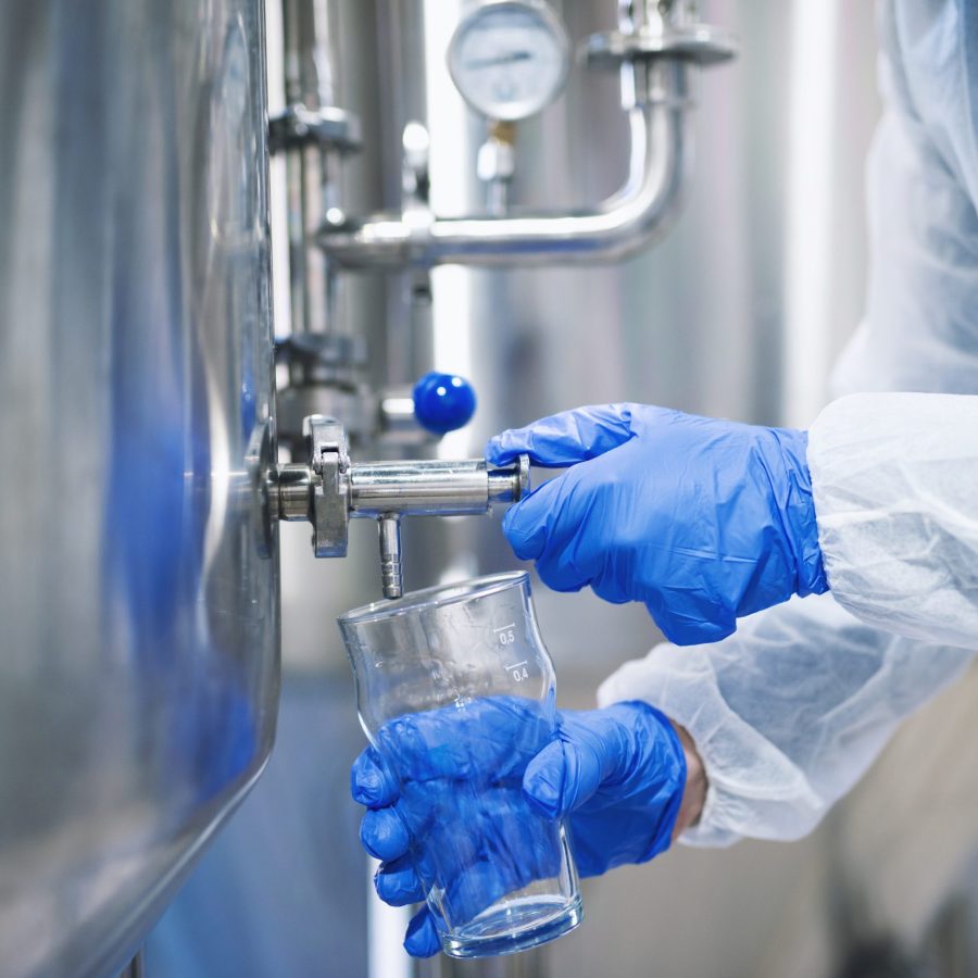 Close up view of hands in protective gloves opening tap valve and filling glass with samples from industrial machine reservoir.